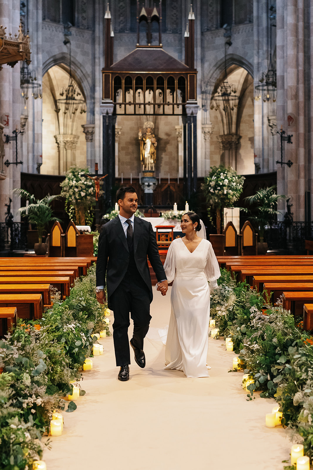 Los novios pasean hacia la salida de la Basílica San Vicente Ferrer de Valencia por un pasillo decorado con verdes.